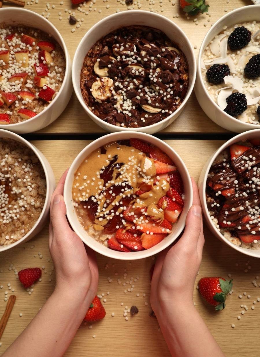 Six bowls of oatmeal and yogurt topped with different fruits and LIFTER Crispies, with hands holding one bowl in front